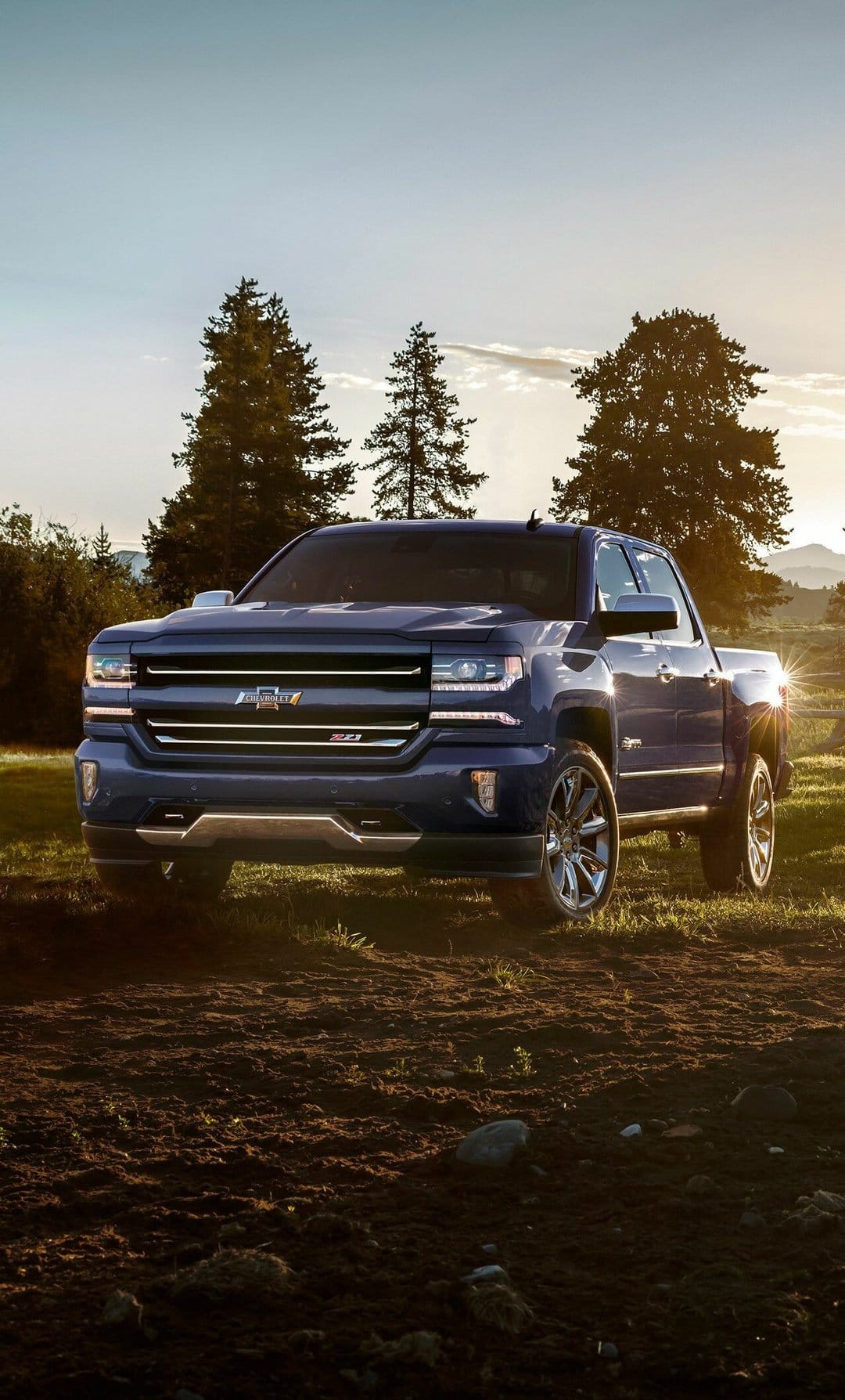Blue Chevrolet Silverado pickup truck in a dirt field at sunset with a lens flare and dark evergreen trees.