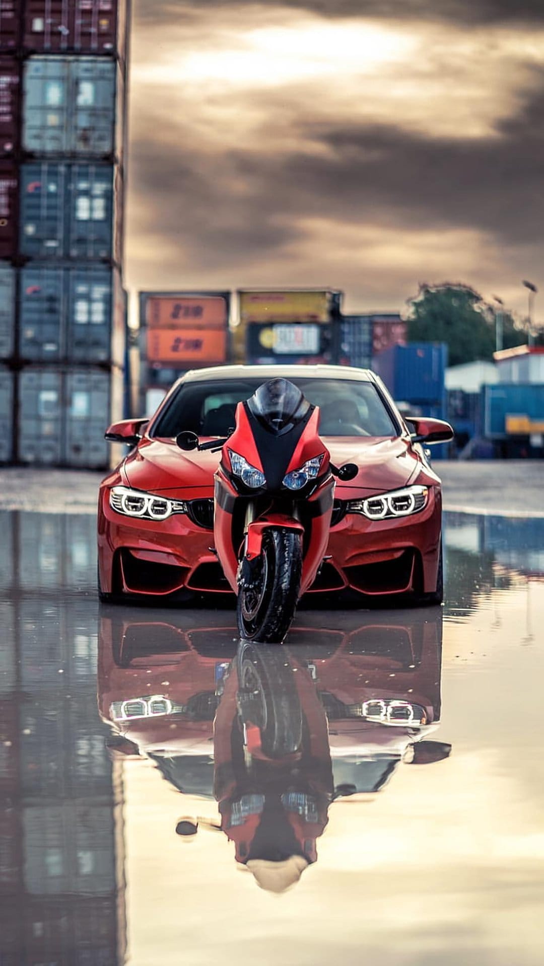 Scarlet sports car and motorcycle parked by shipping containers with symmetrical reflections on wet pavement.