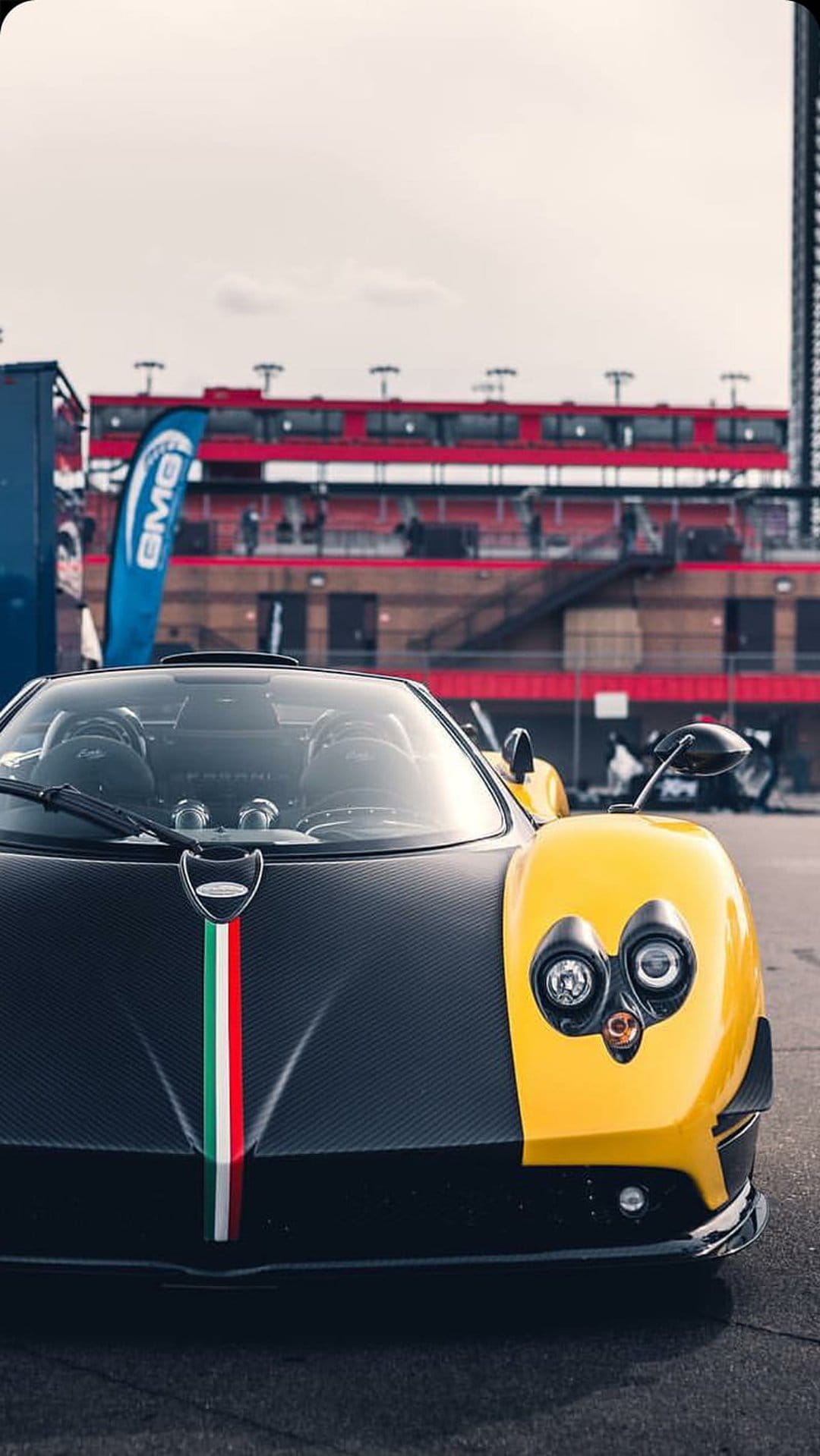 Yellow Pagani supercar with carbon fiber bodywork and Italian flag stripe at a race track under daylight.