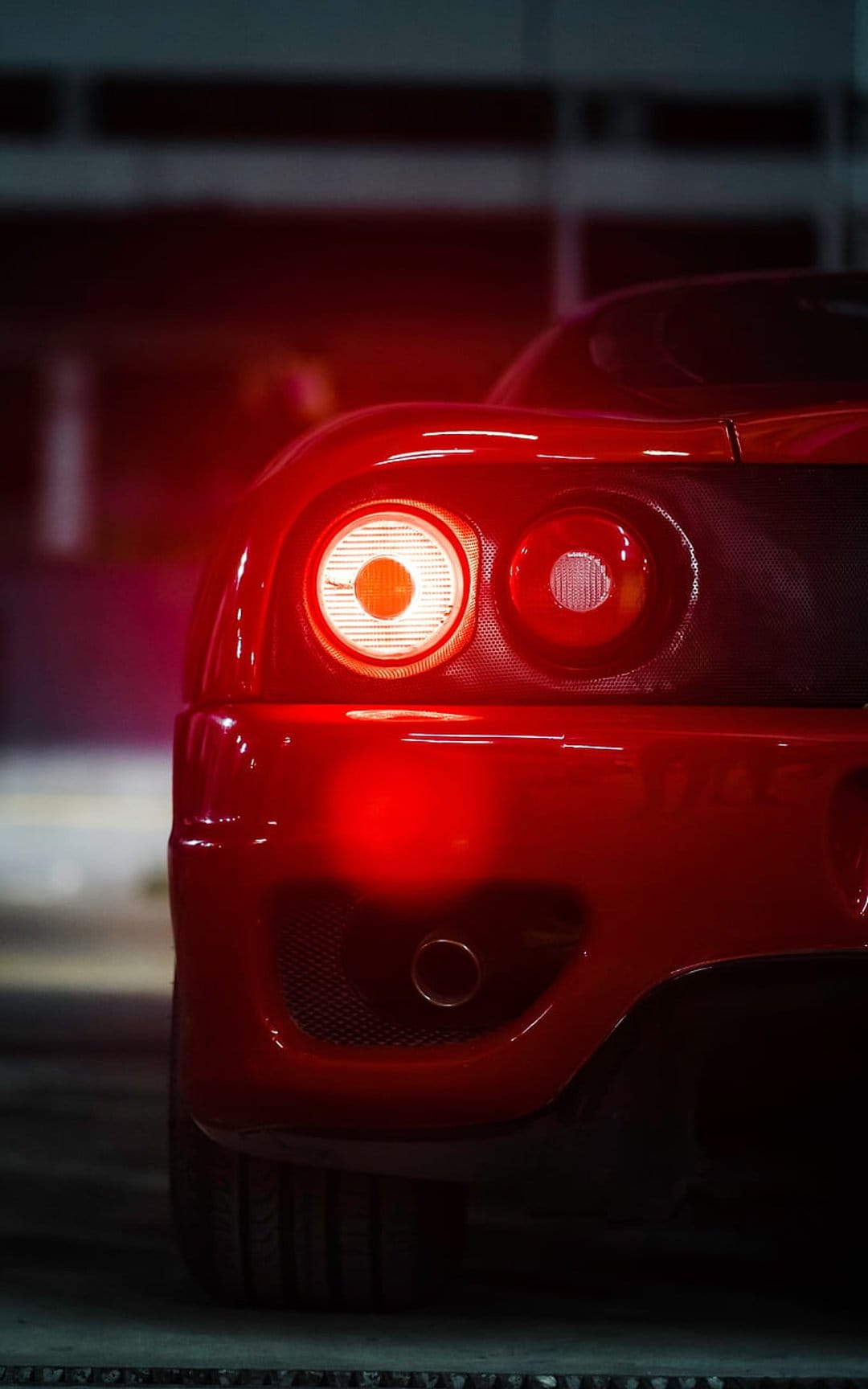 Red Ferrari circular tail lights illuminate a sleek car body with a warm crimson glow in a tight close-up.