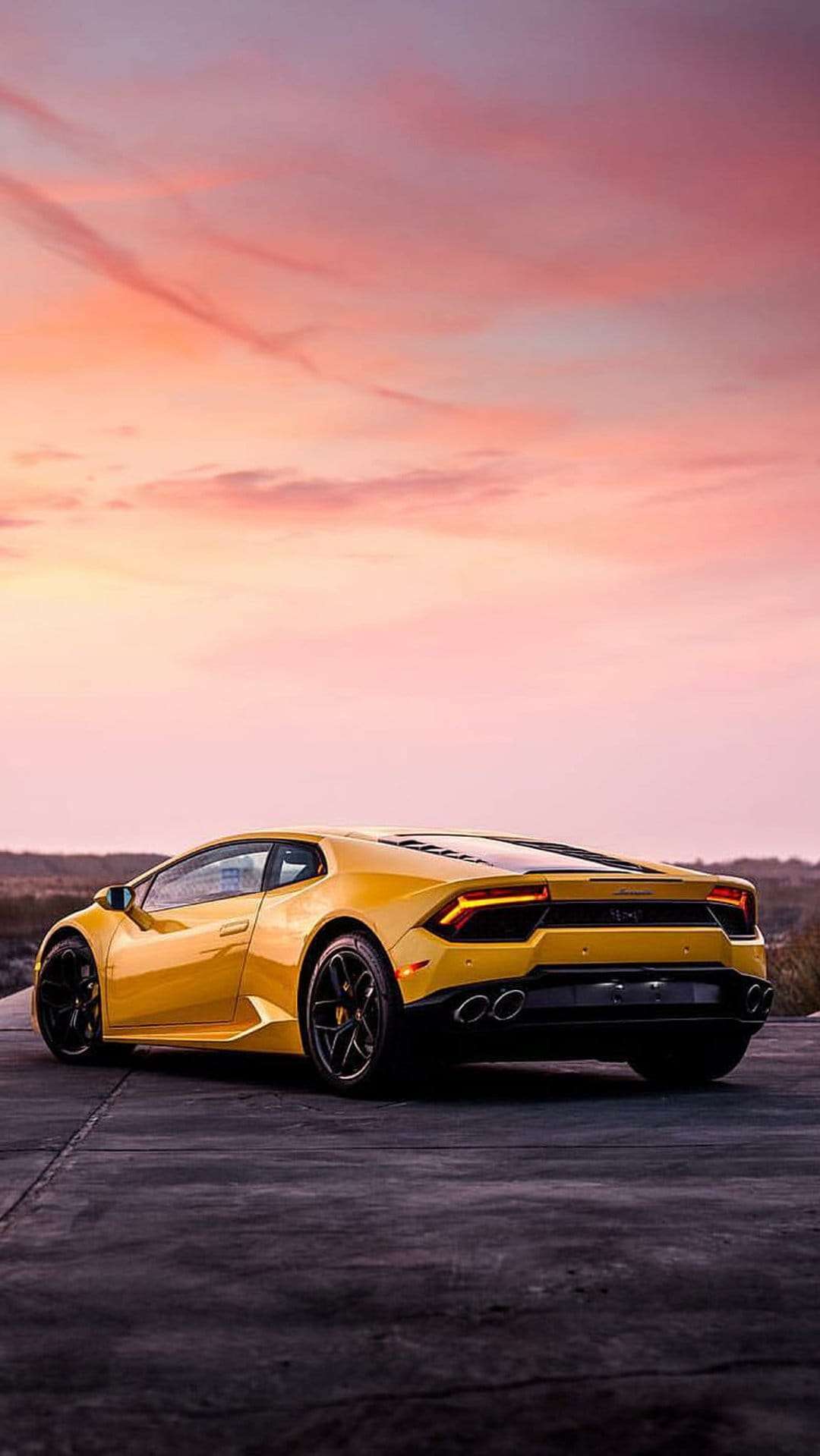 Yellow Lamborghini Huracan parked on asphalt during a vivid pink and orange sunset with dramatic cloud formations.