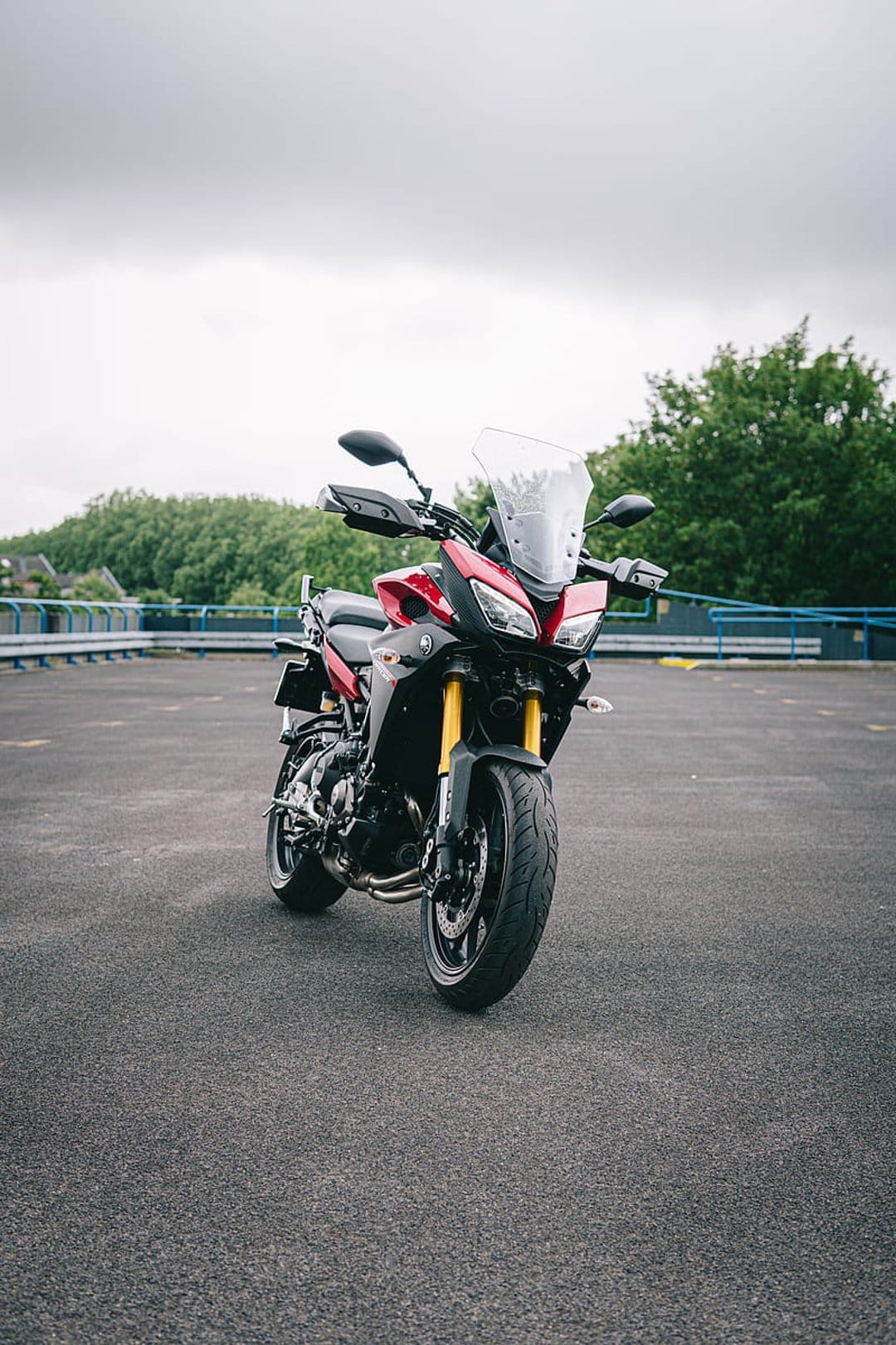 Red Yamaha motorcycle with yellow forks parked on asphalt against lush green trees and a cloudy gray sky.