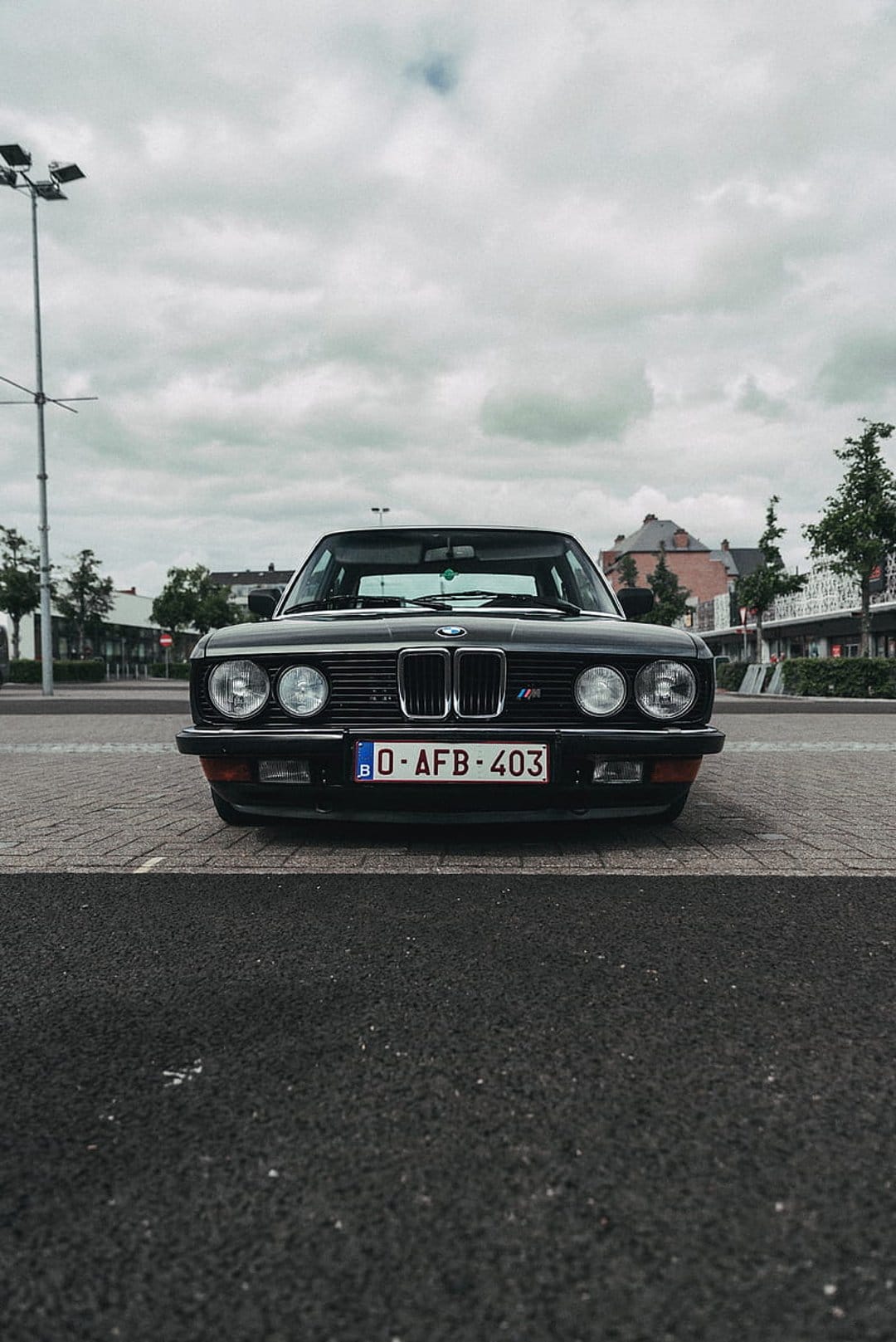 BMW E28 M535i sedan with a black finish and classic round headlights parked on a gray cobblestone street.