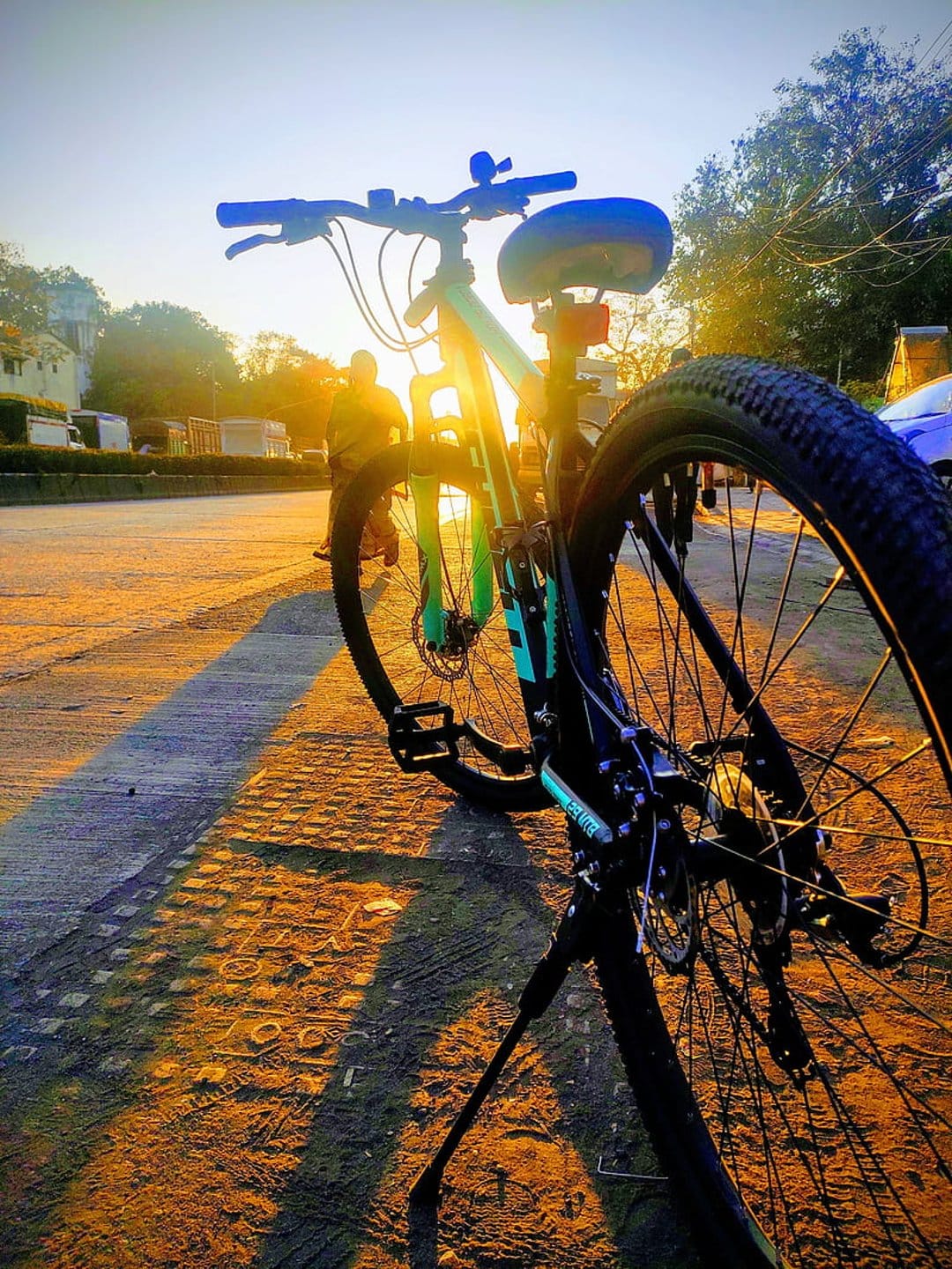 Bicycle parked on a dirt trail at sunset with long shadows and a distant cyclist under a glowing golden sky.
