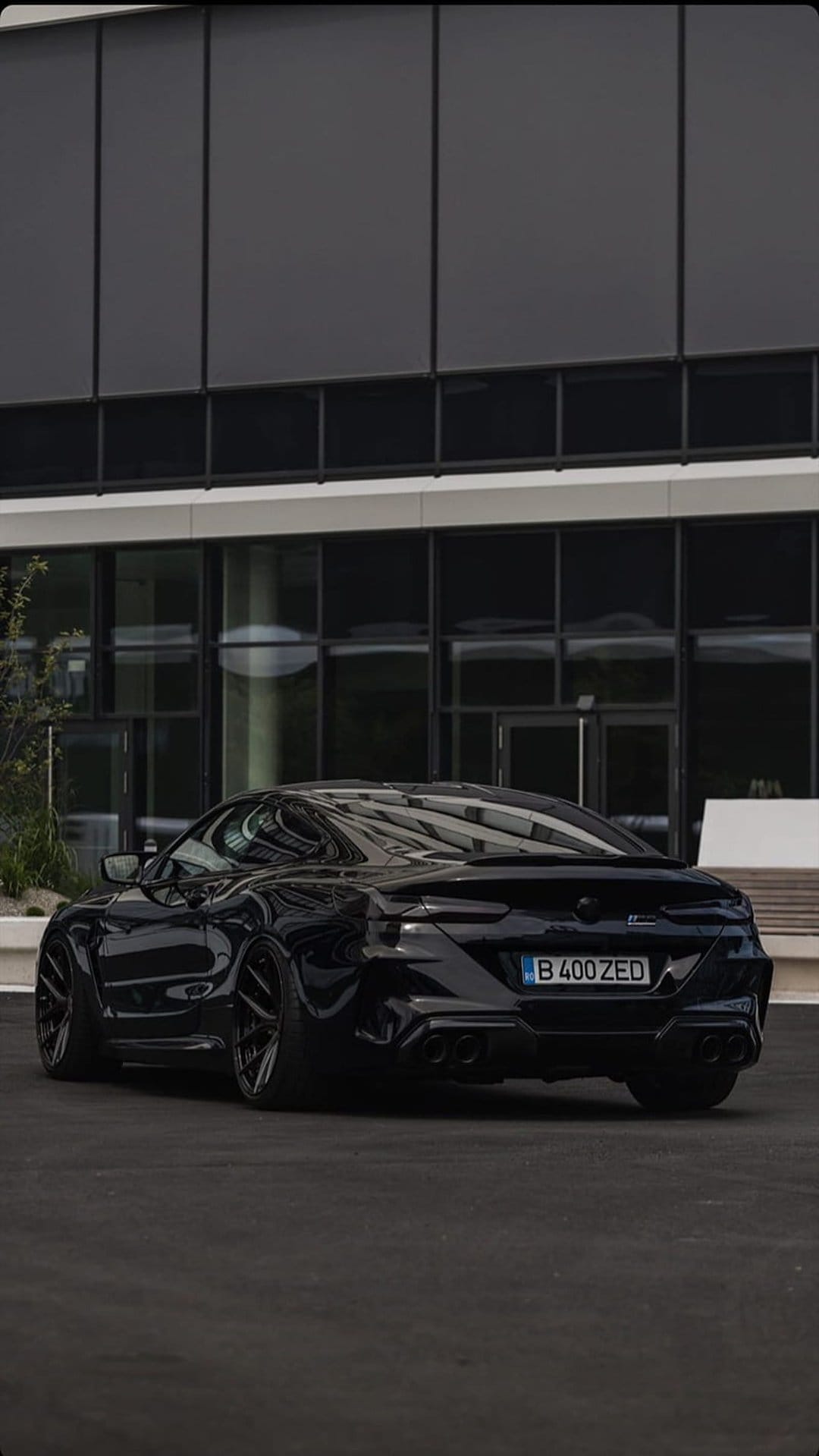 Black sports car with widebody fenders parked on asphalt in front of a modern glass building at night.