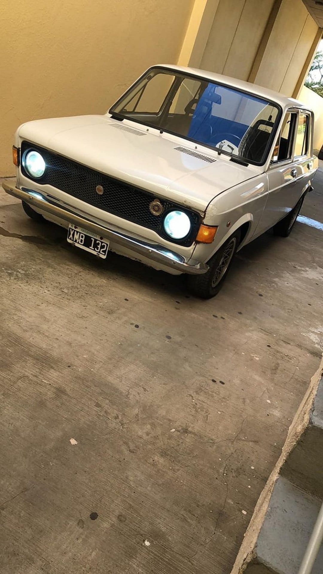Fiat 128 sedan in white parked by a yellow wall with chrome bumpers and classic headlights on a concrete floor.