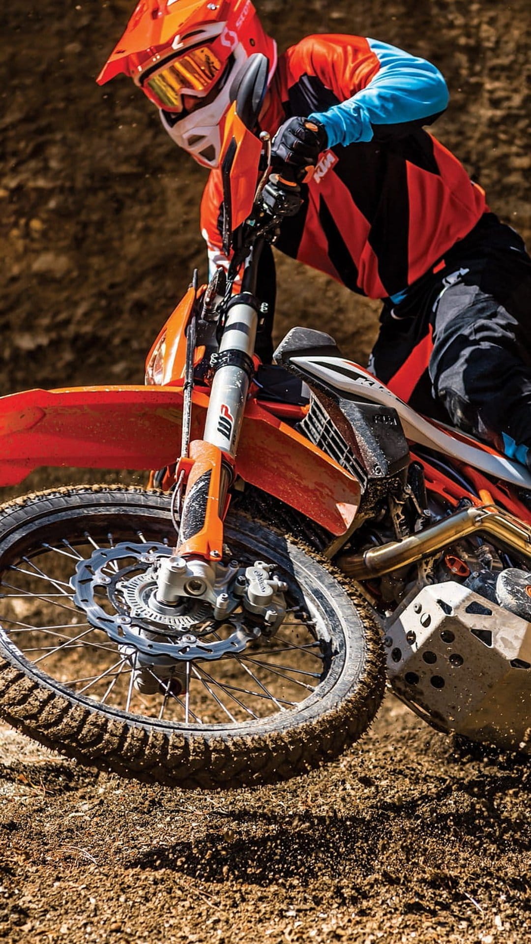 Motocross rider in orange gear leans a dirt bike into a sharp turn on a dusty brown track with front wheel airborne.