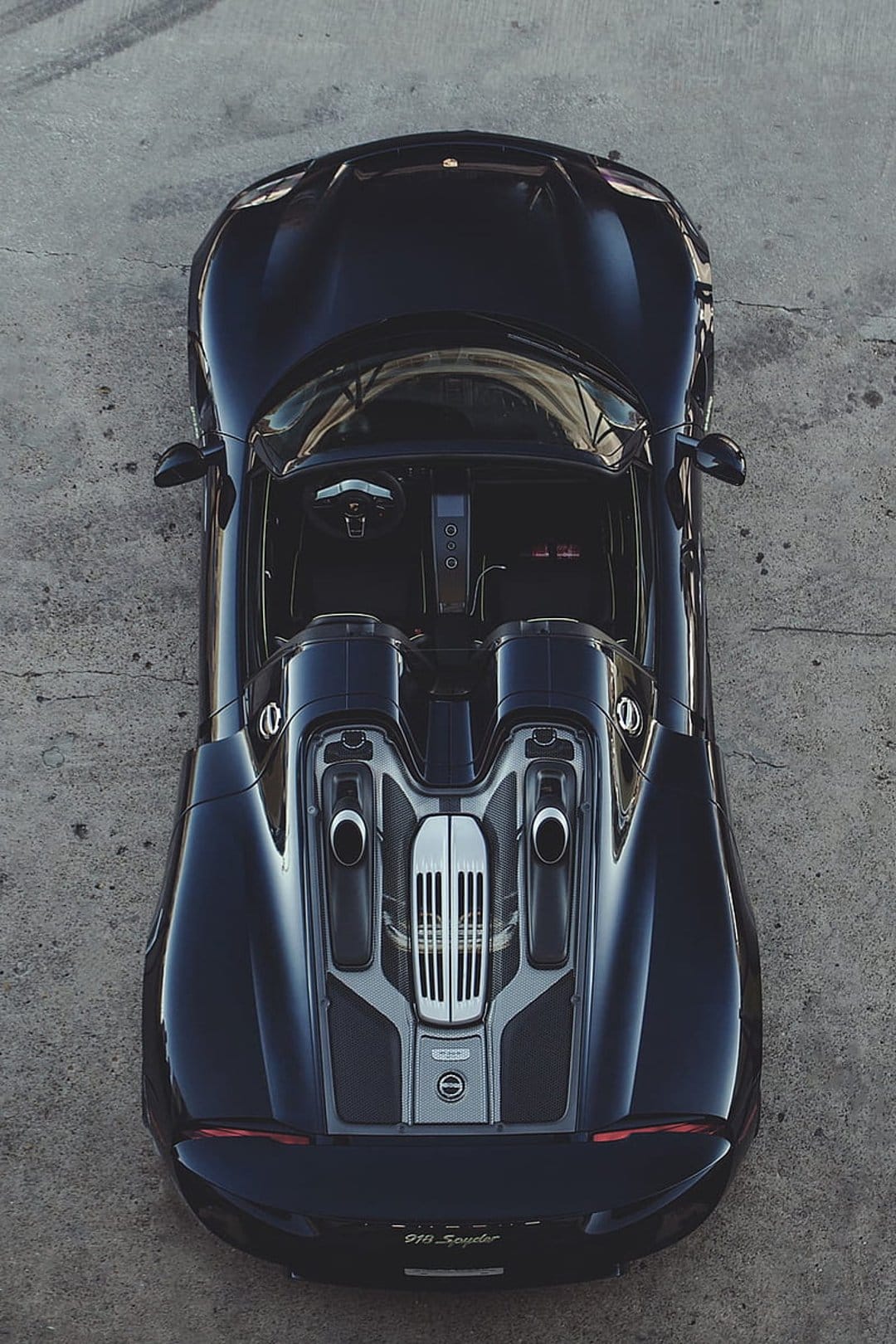 Porsche 918 Spyder in black seen from above on a gray concrete floor with visible dual exhaust pipes.