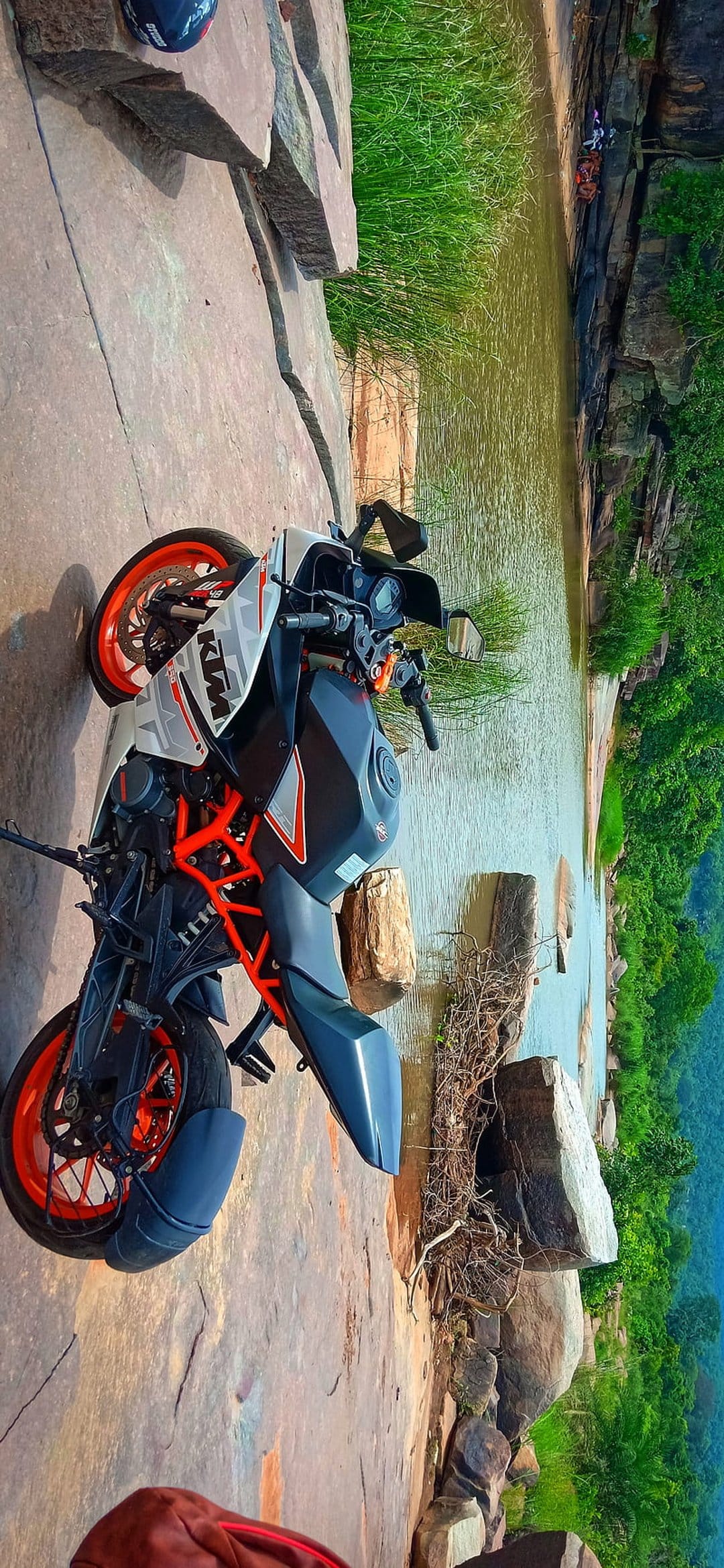 KTM RC sports bike parked on a flat rocky outcrop overlooking a lush riverbank with dense green vegetation.