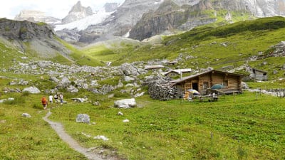 Hikers on a dirt path lead toward a rustic wood cabin at the base of snowy Swiss mountain peaks under a gray sky.