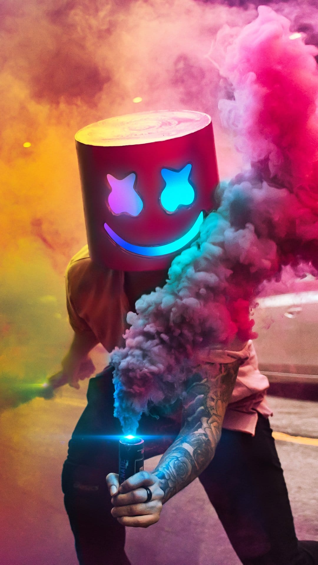 Marshmello in a glowing helmet holds a pink smoke bomb, showing a tattooed arm against a dark urban street.