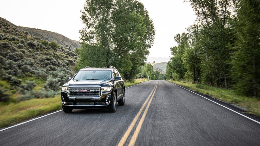 Black GMC SUV driving on a paved road with double yellow lines between green trees and a grassy hillside.