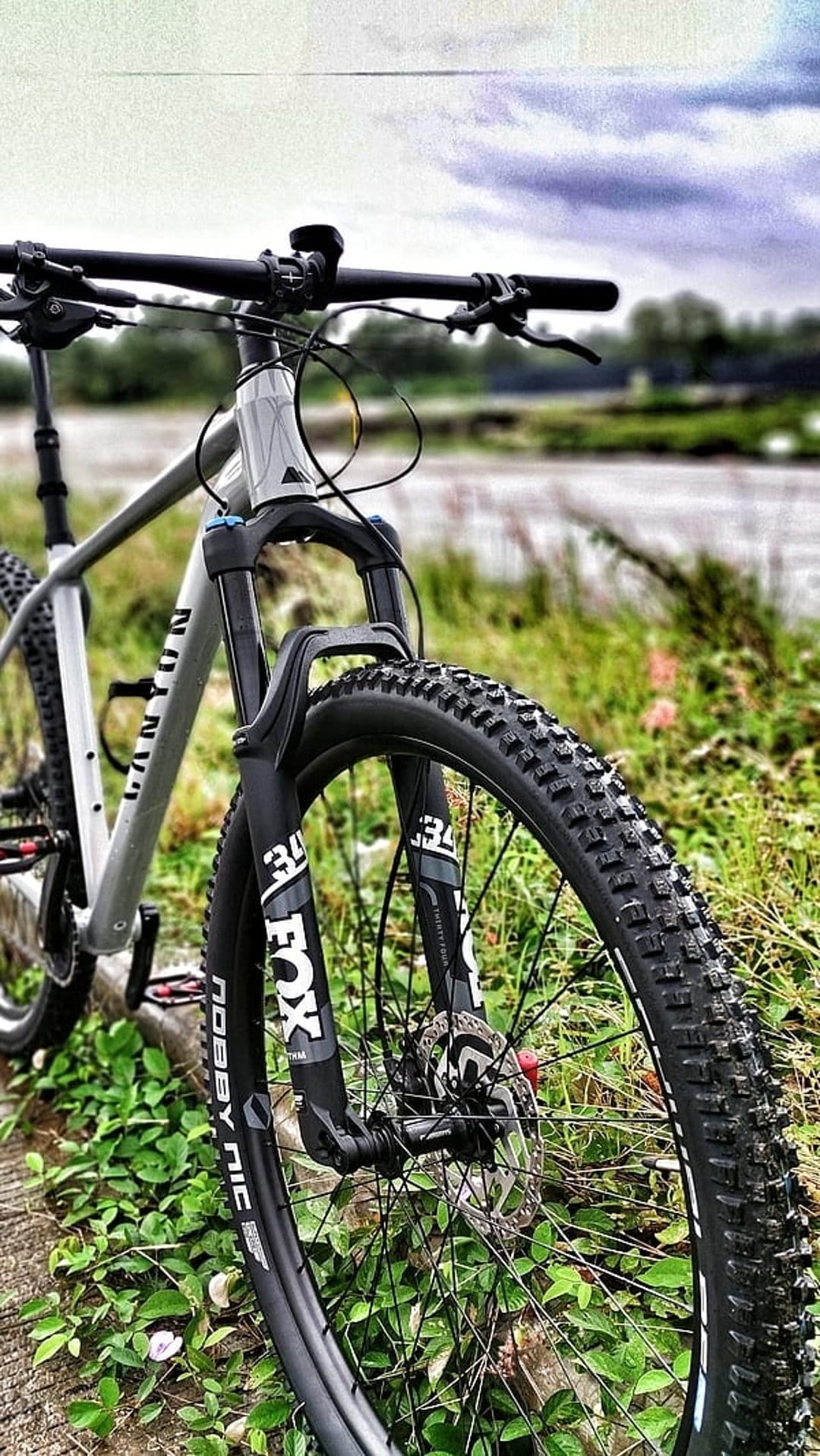 Canyon mountain bike front suspension and knobby tires parked on a riverbank under a cloudy grey sky.