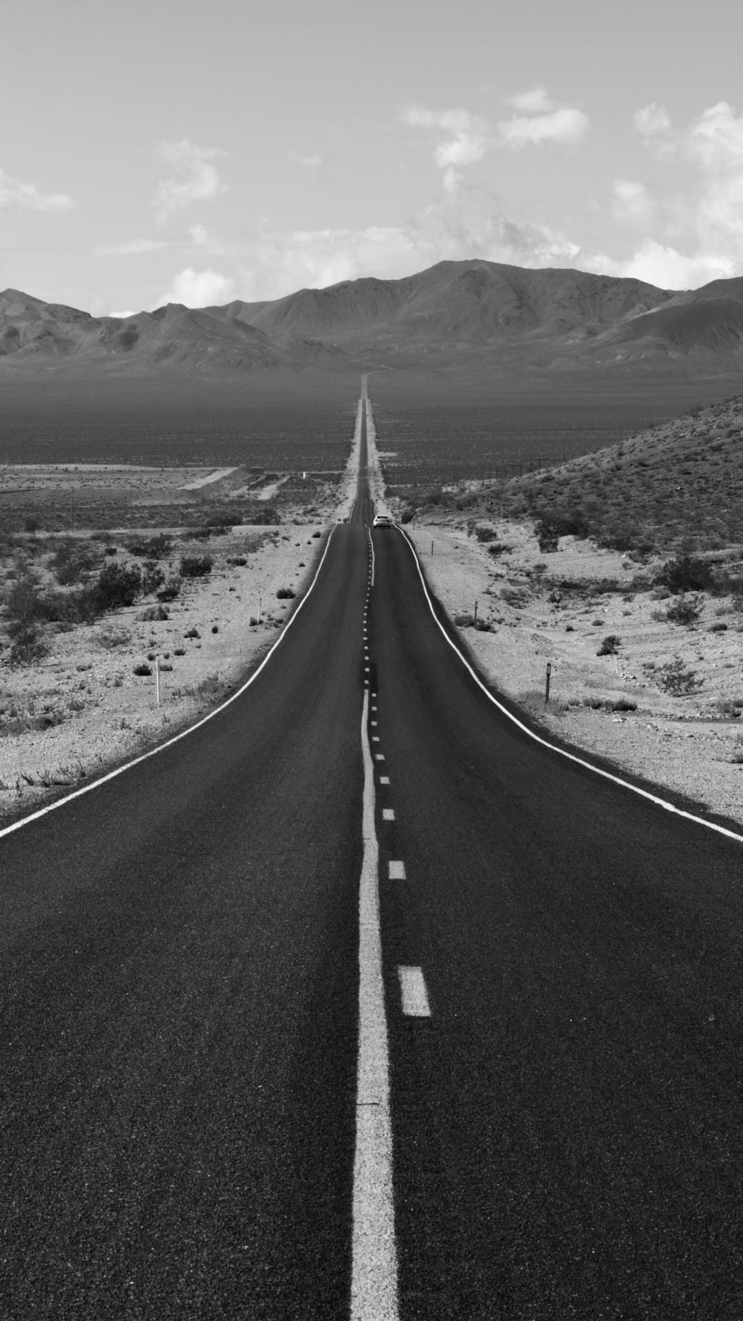 A straight highway recedes into the distance between rugged hills under a clear sky in black and white.