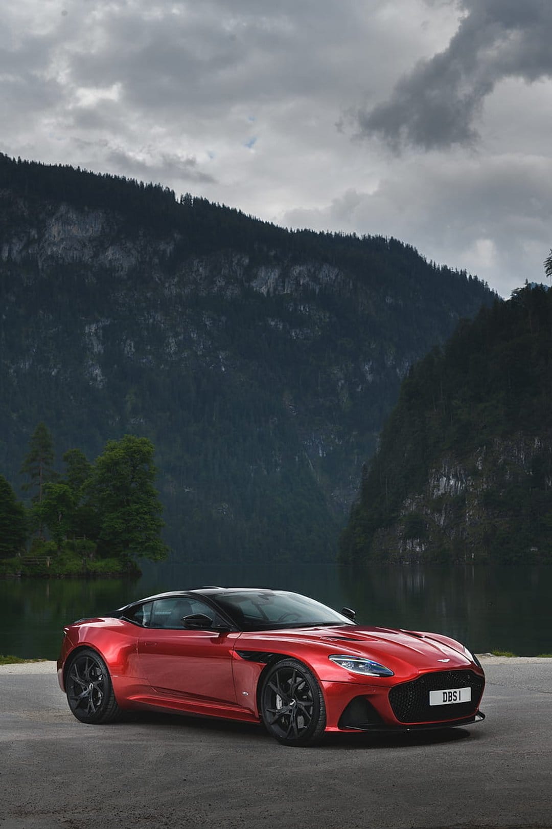 Red Aston Martin DBS Superleggera parked on a mountain road beside a calm lake beneath a dramatic cloudy sky.