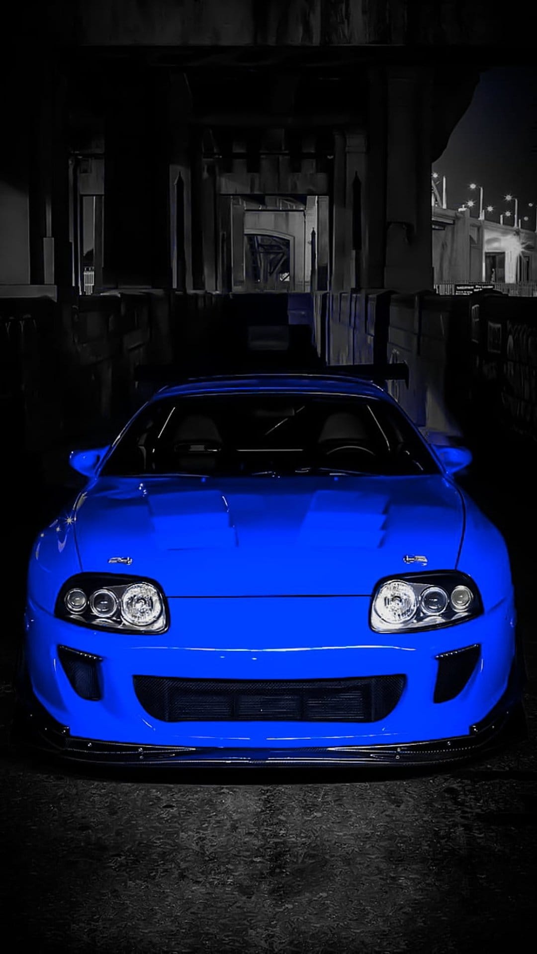 Blue Toyota Supra with glowing headlights parked in a dark industrial underpass from a low-angle perspective.