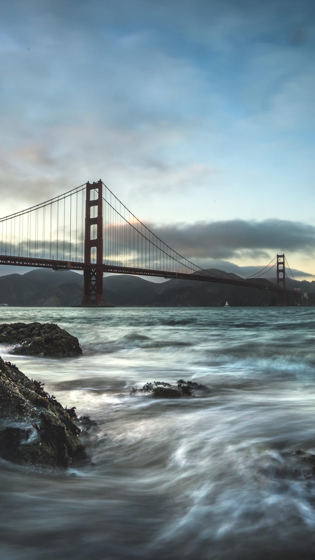 Golden Gate Bridge towers over crashing waves and jagged rocks under a misty twilight sky in San Francisco.