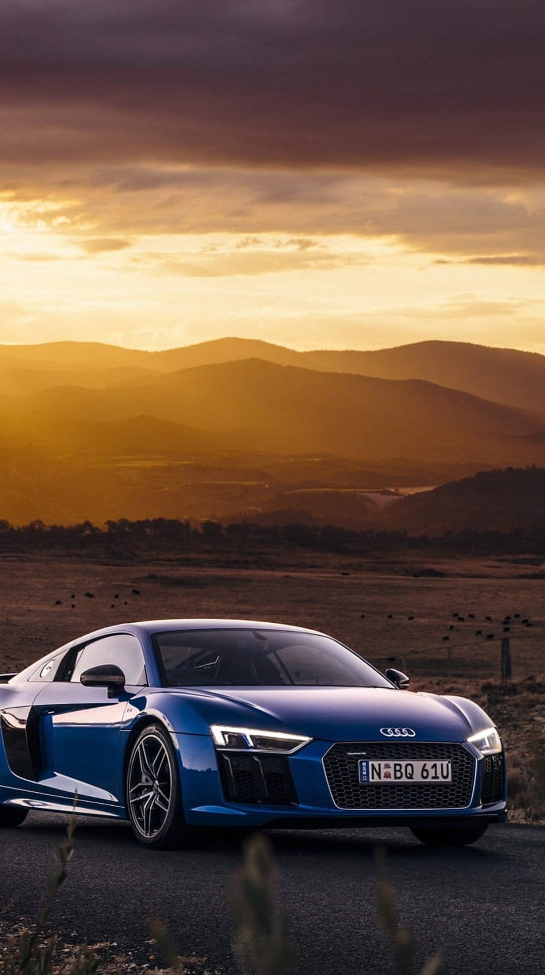 Blue Audi R8 sports car driving on a mountain road during a golden sunset with dramatic clouds and peaks behind.