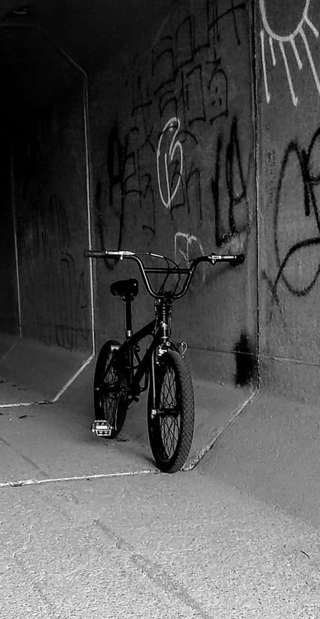 Black BMX bicycle stands against a wall of layered graffiti inside a dark monochromatic concrete tunnel.