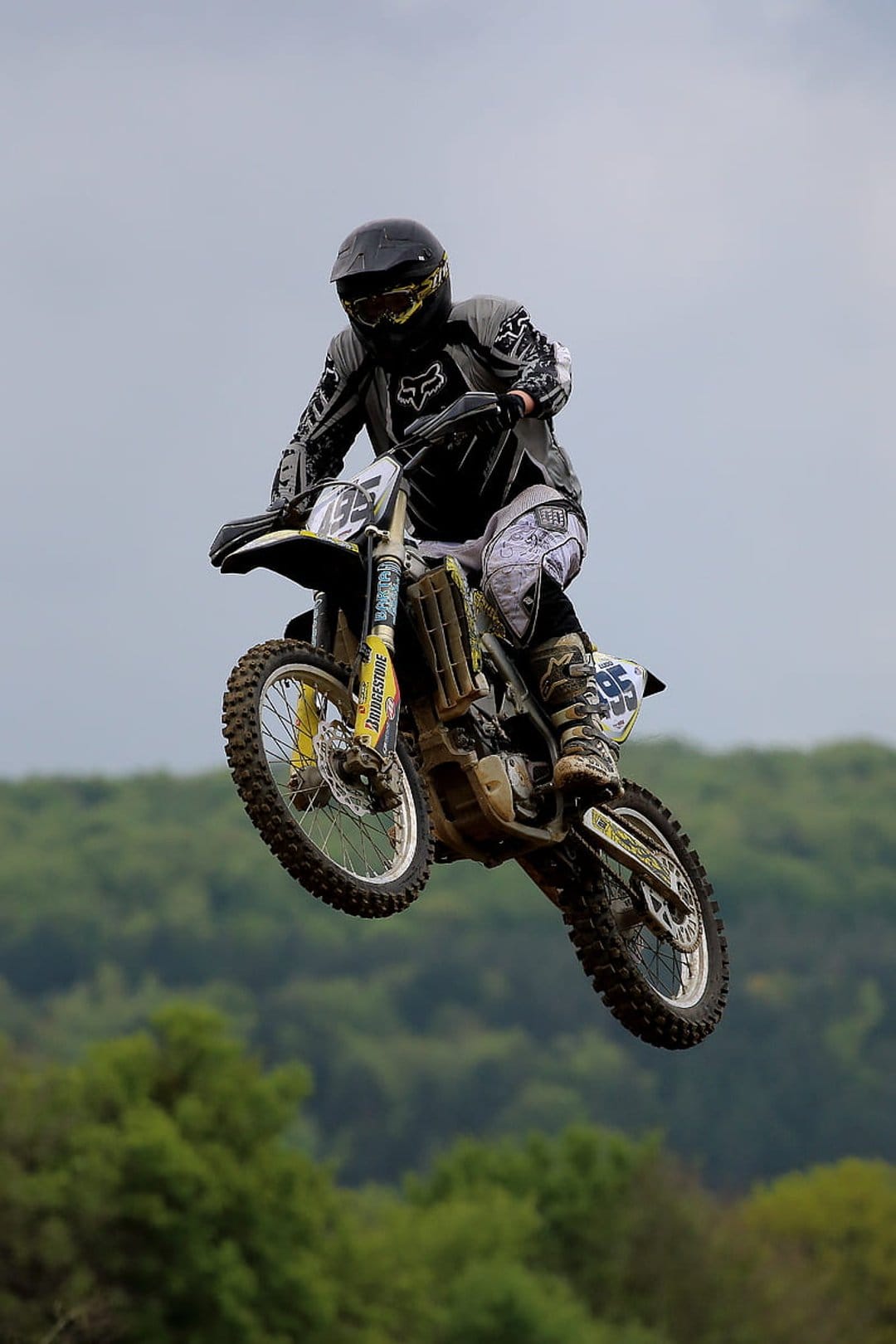 Motocross rider in full protective gear jumping a dirt bike high against an overcast sky and blurred treeline.