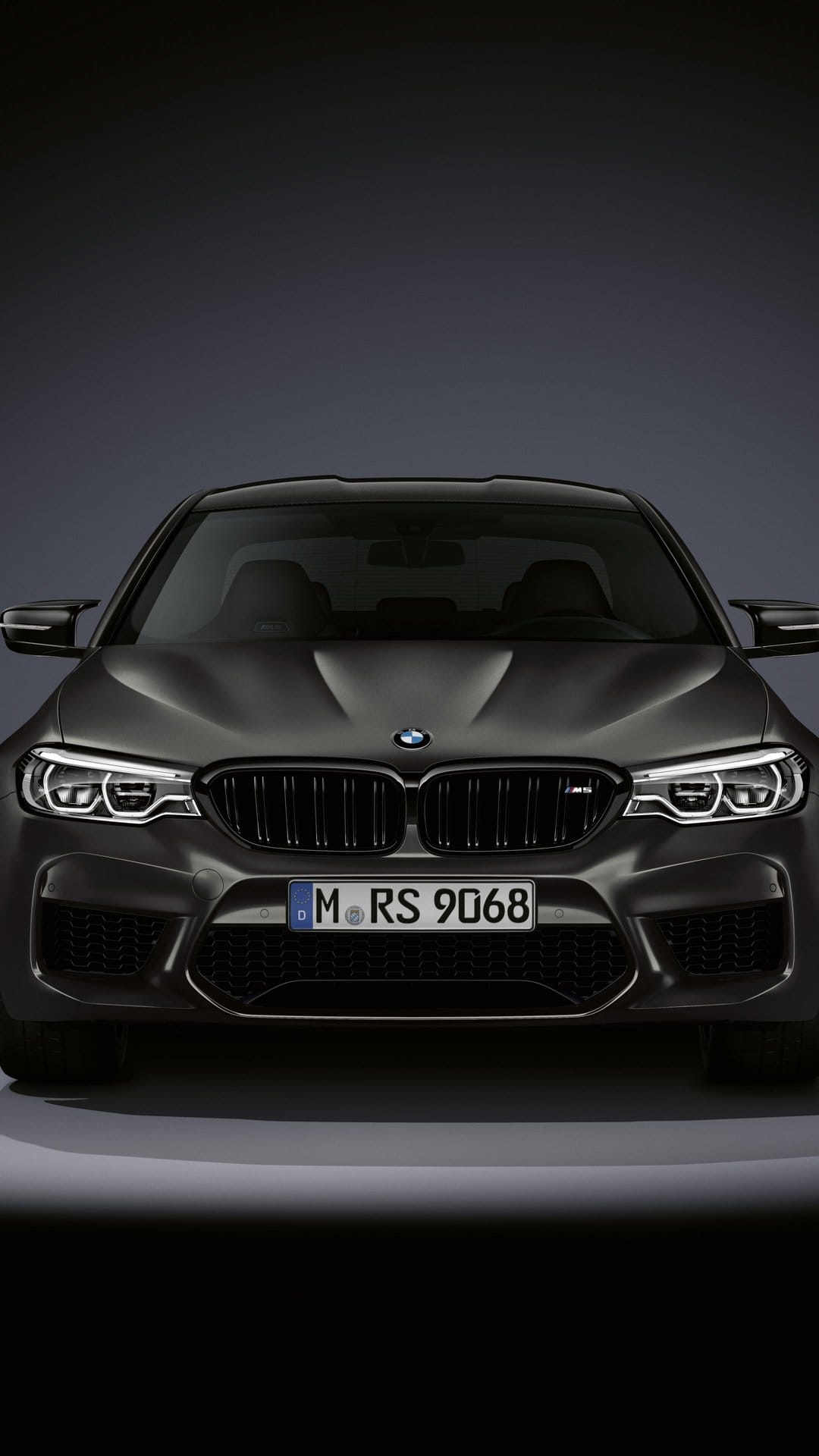 Front view of a dark matte grey BMW M5 with glowing LED headlights and a German license plate on a dark background.