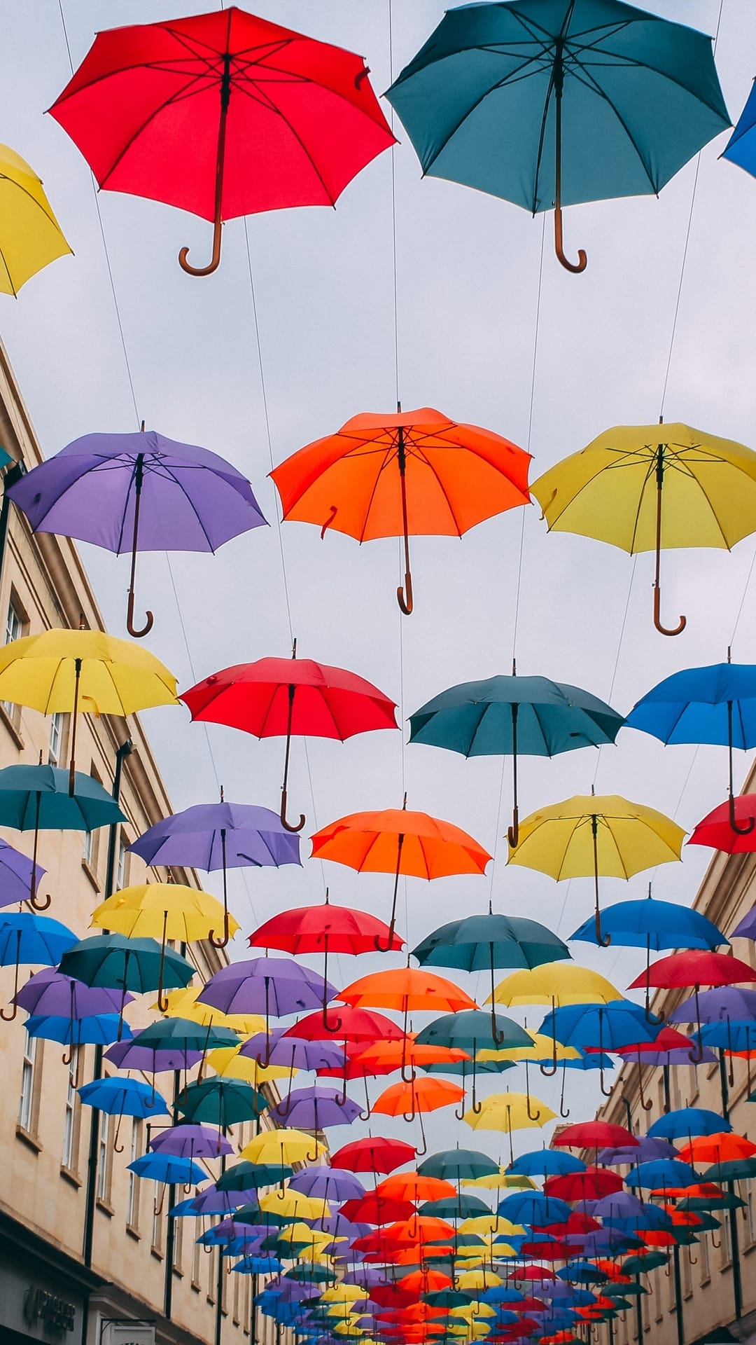 Multi-colored umbrellas suspended over a narrow street under a soft gray sky with vibrant ambient lighting.