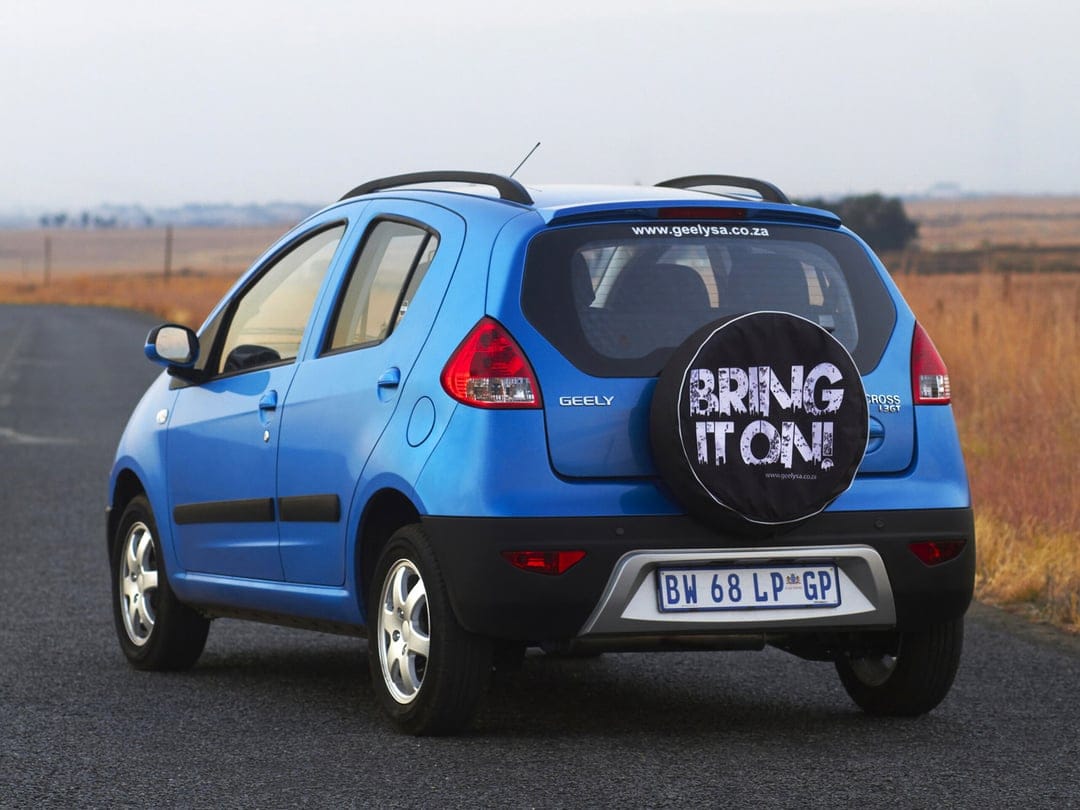 Blue Geely LC Cross hatchback parked on a quiet asphalt road with a spare tire cover and golden fields.