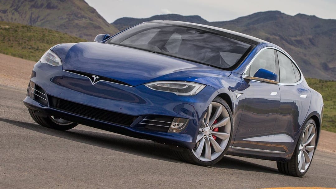 Blue Tesla Model S with silver wheels and red calipers parked on an asphalt road against mountain peaks.