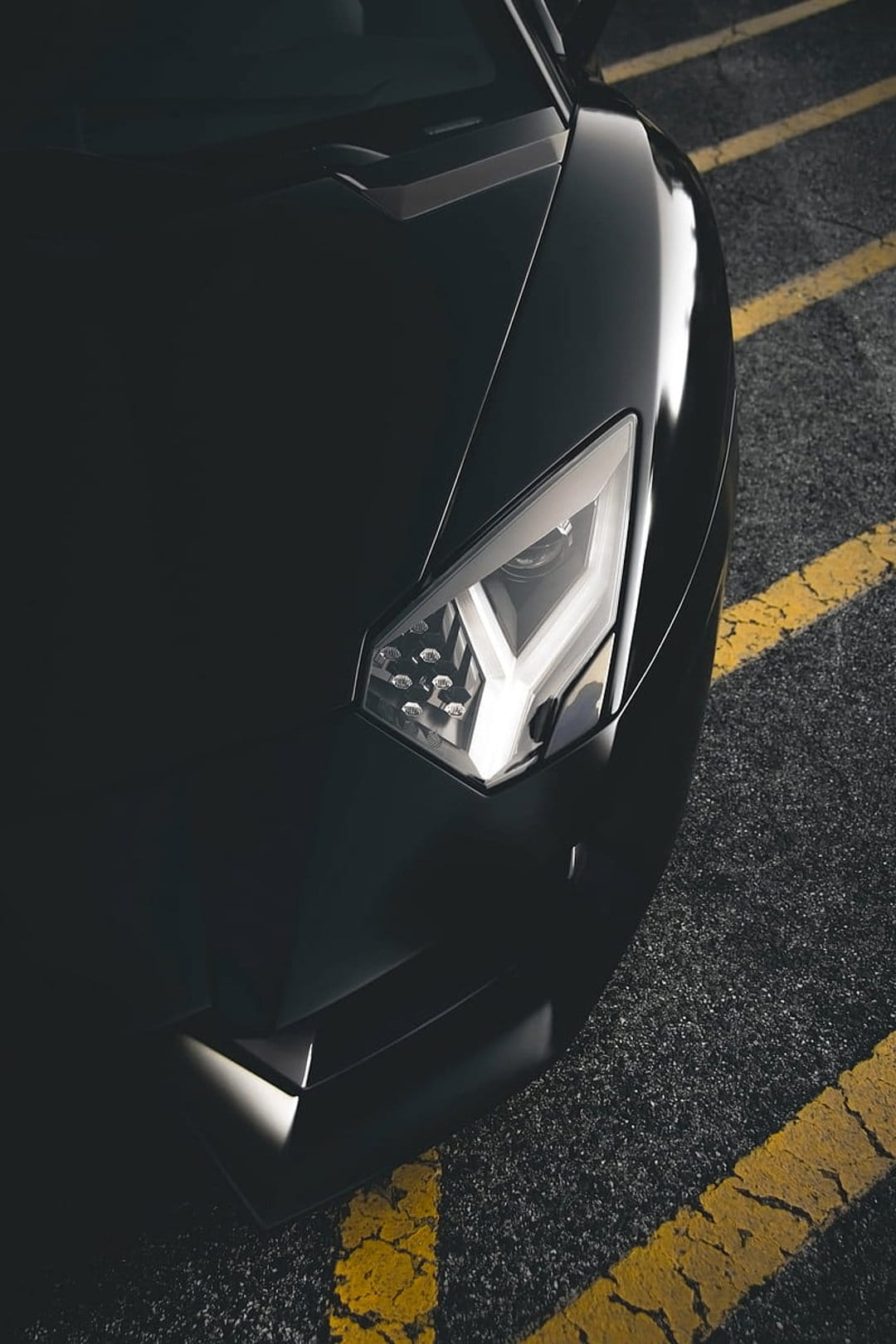 Lamborghini Aventador LED headlight and front bumper over yellow pavement markings on dark weathered asphalt.