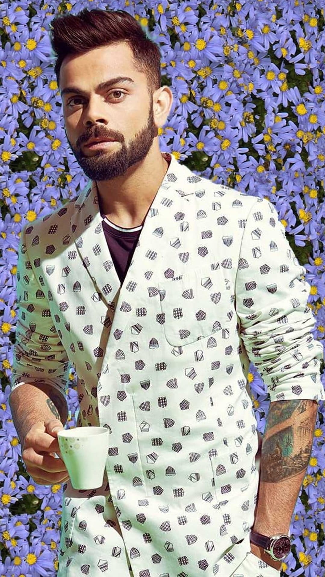 Virat Kohli in a patterned jacket holding a coffee cup before a dense wall of purple and yellow flowers.