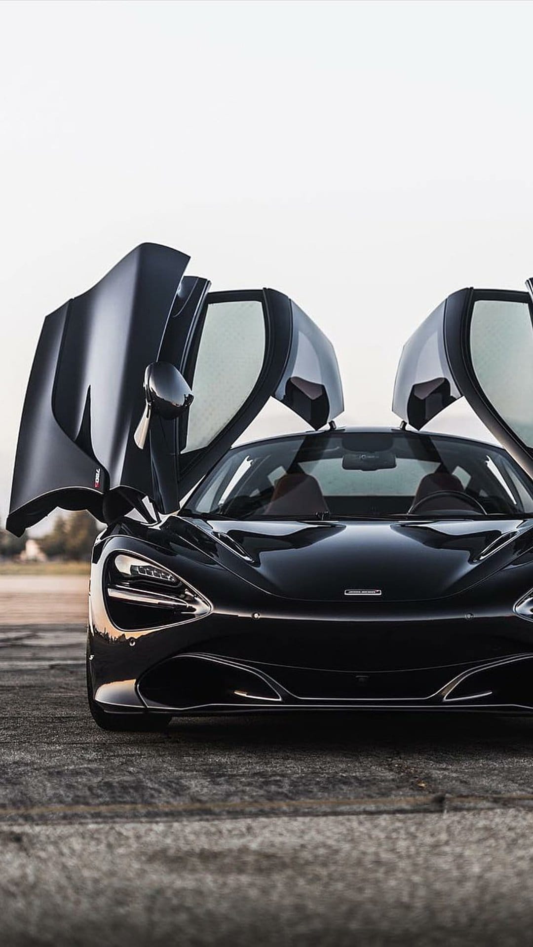 Black McLaren 720S supercar with open dihedral doors on textured asphalt under soft lighting at a low angle.