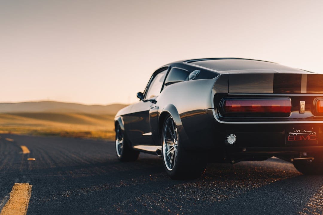Classic dark grey muscle car on a winding road during golden hour with blurred hills in the background.