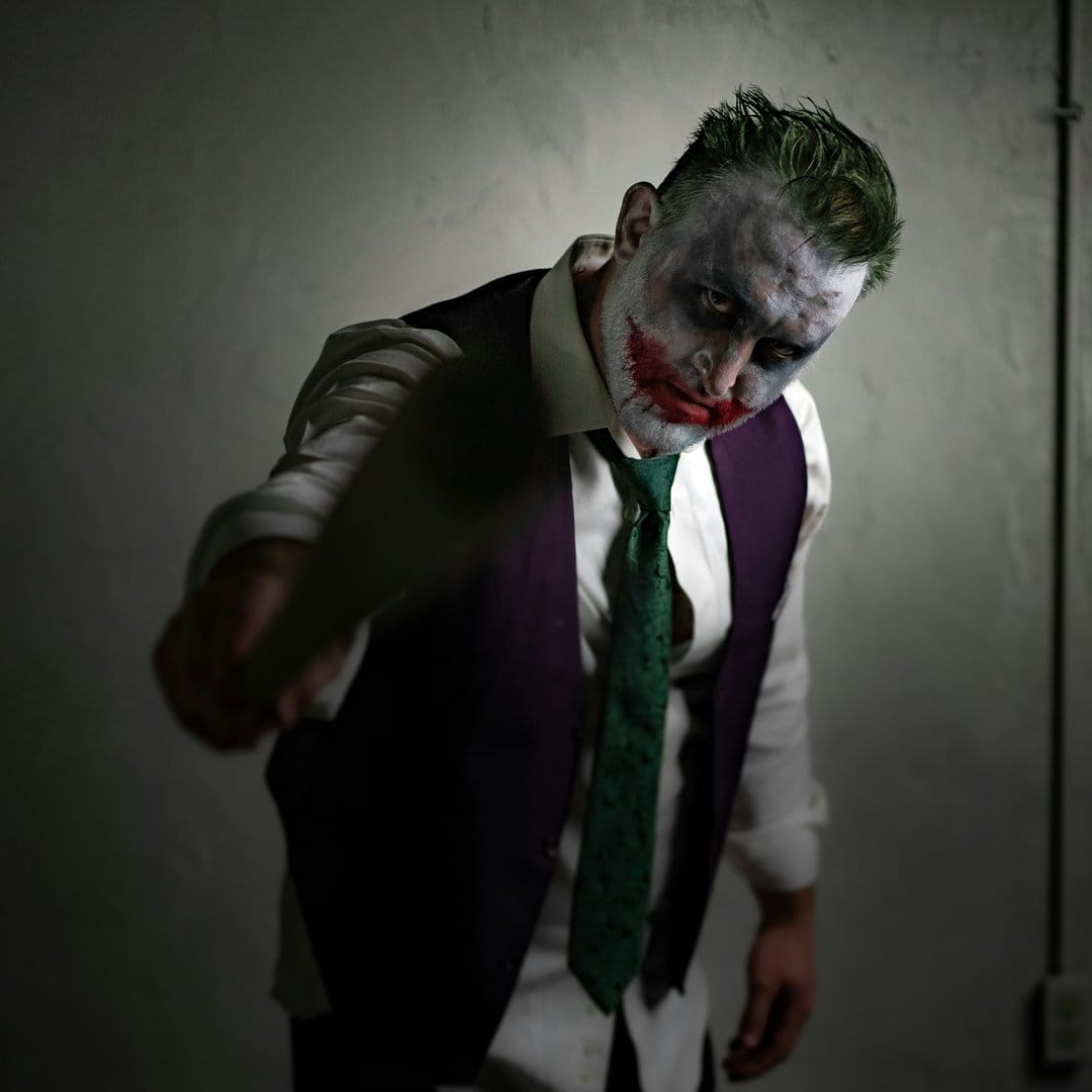 Joker cosplayer with green hair and a red smile holds a knife toward the lens under dramatic low-key lighting.