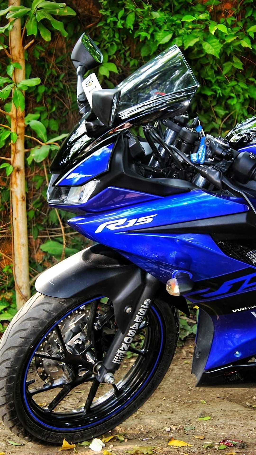 Blue Yamaha R15 sportbike with glossy fairings parked near green leaves showing front wheel and disc brake details.