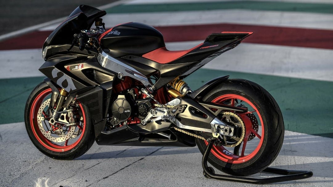 Side view of a carbon fiber Aprilia sportbike with red wheels and seat on a racetrack with Italian flag colors.