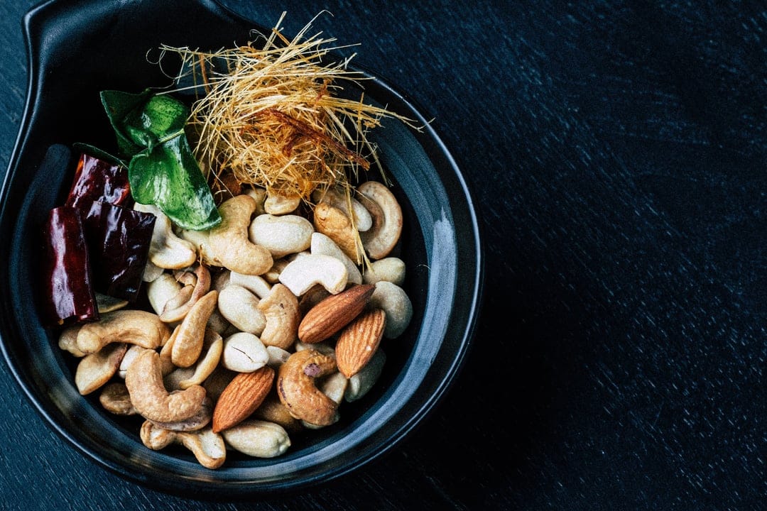 Cashews and almonds mixed with red chilies and herbs in a dark bowl, viewed from a top-down perspective.