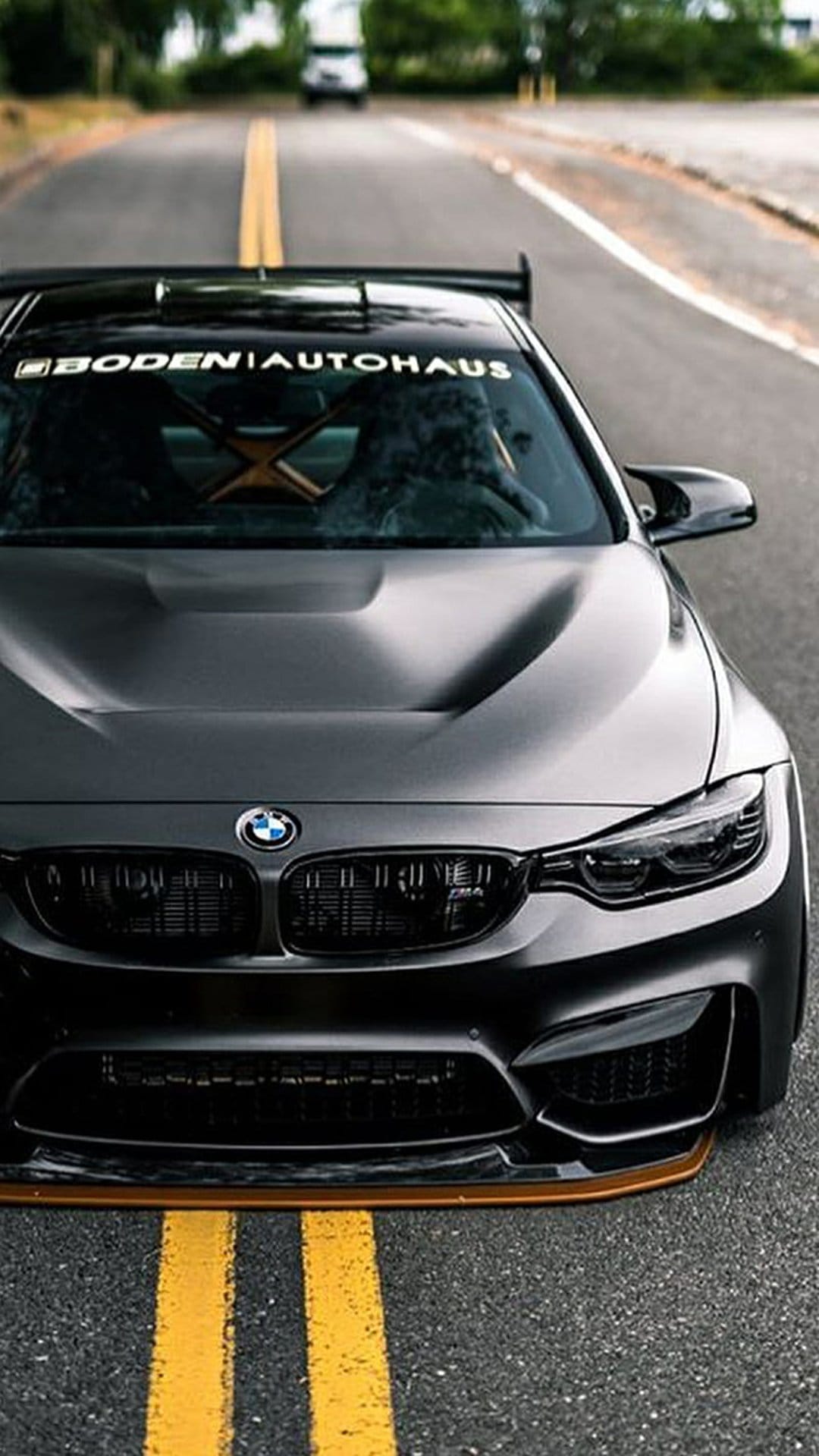 Dark BMW M4 with a large spoiler and windshield decal sits on an asphalt road with yellow dividing lines.