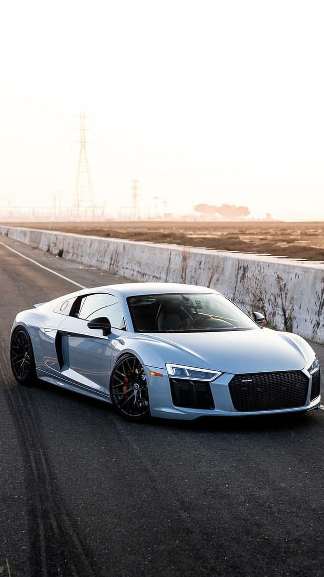 Silver Audi R8 sports car parked on a paved road with tire marks against a hazy sunset and industrial horizon.