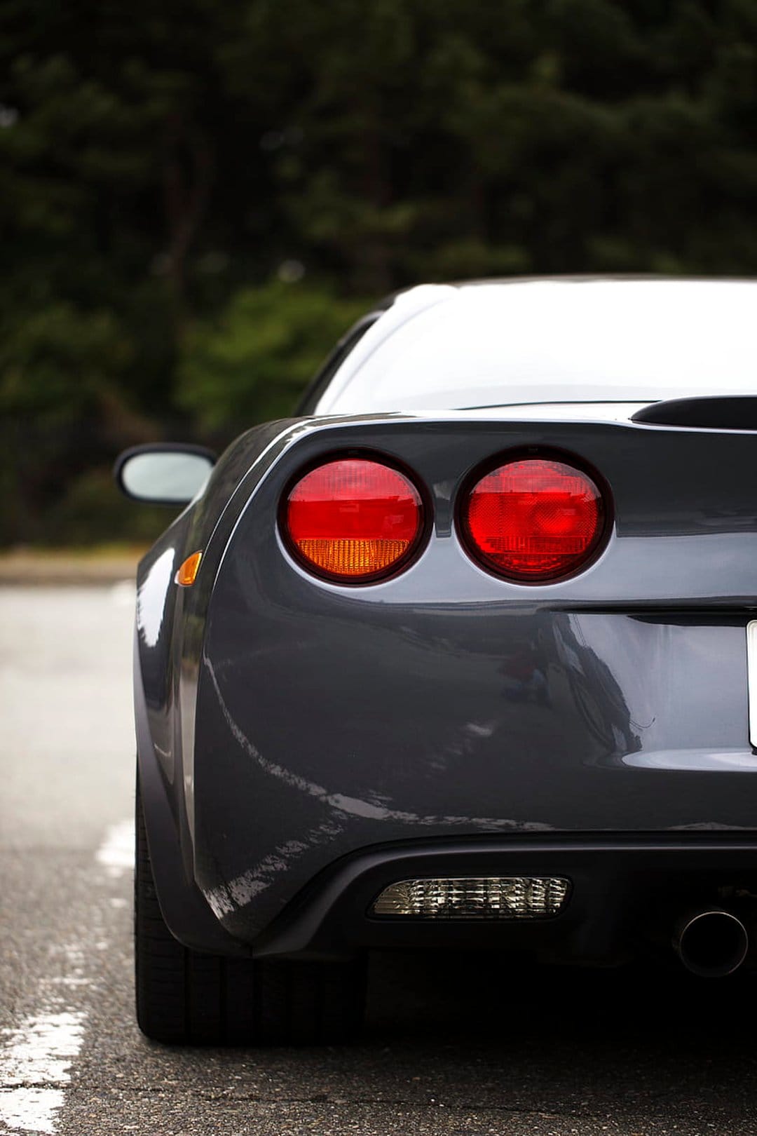 Gray Chevrolet Corvette rear bumper and circular taillights with glossy paint reflections on a paved road.