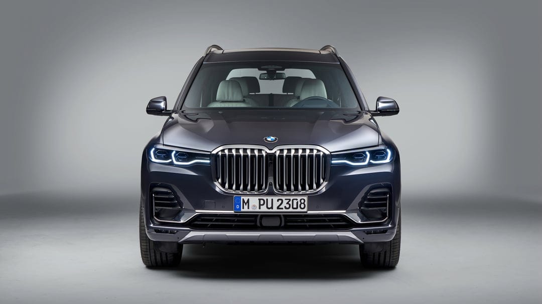Dark grey BMW X7 SUV with glowing LED headlights and a large chrome grille in a minimalist studio setting.