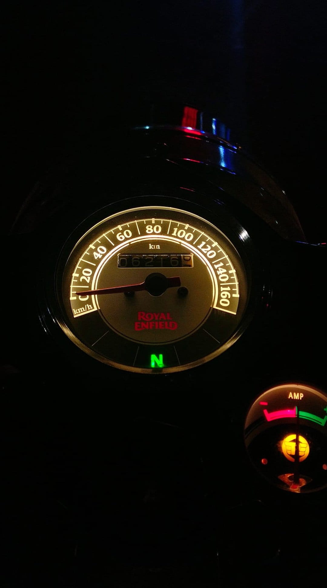 Royal Enfield motorcycle speedometer and amp gauge glowing with green and yellow light in a dark night setting.