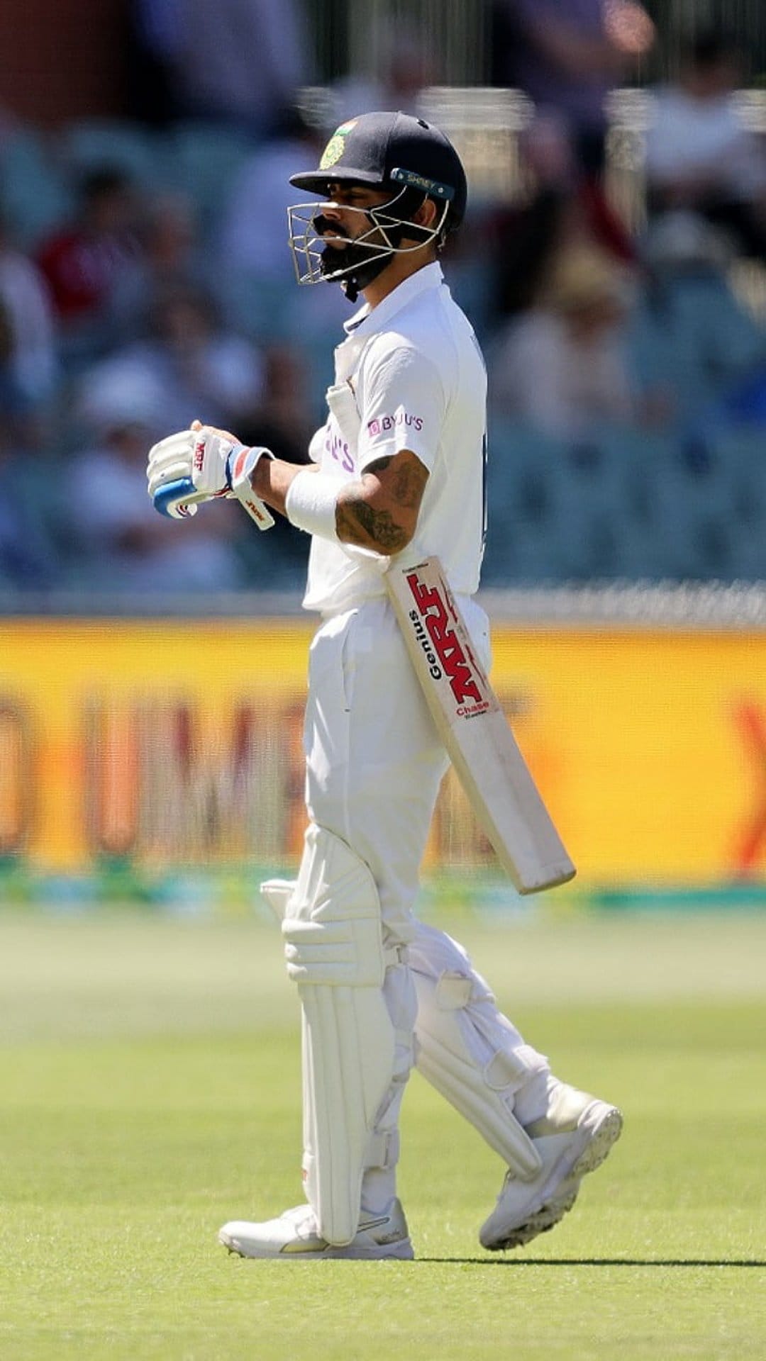 Cricketer in white uniform holding an MRF bat is shown in a side profile view with a blurred stadium backdrop.