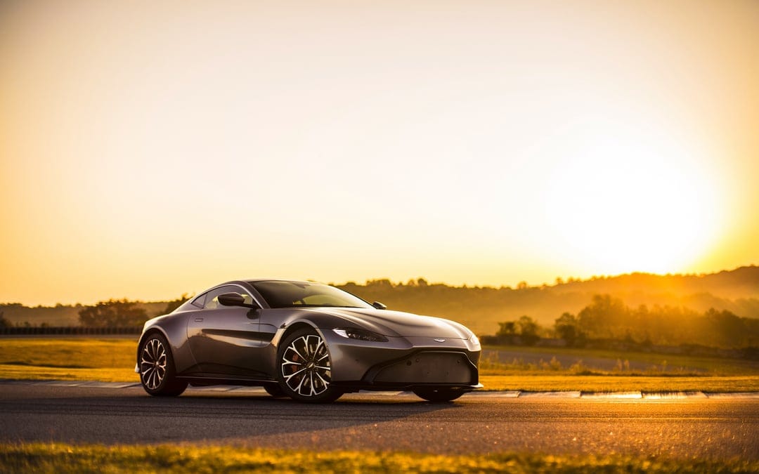 Silver Aston Martin Vantage parked on a racetrack during a warm golden hour sunset with bright sun flare.