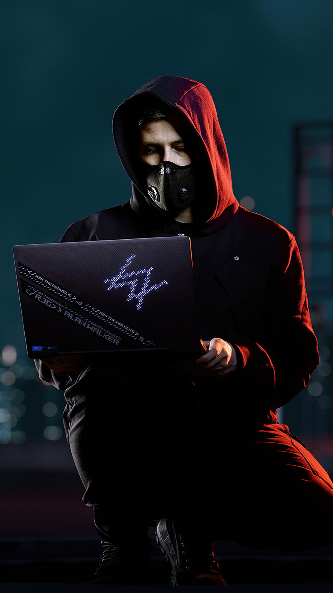 Alan Walker in a black mask and hoodie holds a glowing ROG laptop with a neon logo against a city dusk backdrop.