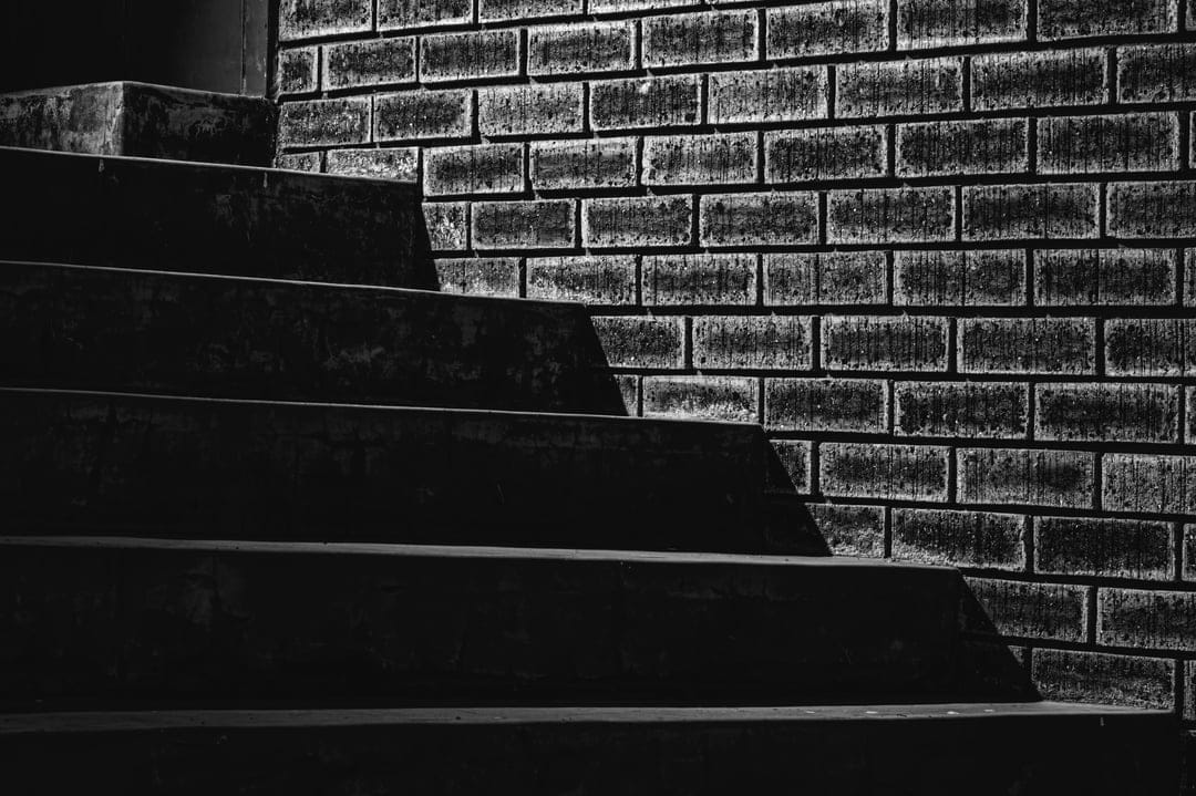 Dark wooden stair treads rise against a weathered brick wall in a high-contrast black and white composition.