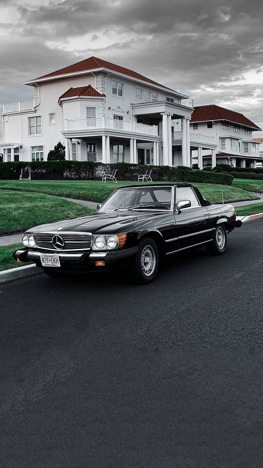 Black Mercedes SL convertible parked on a driveway in front of a white mansion with a red tile roof and lawn.