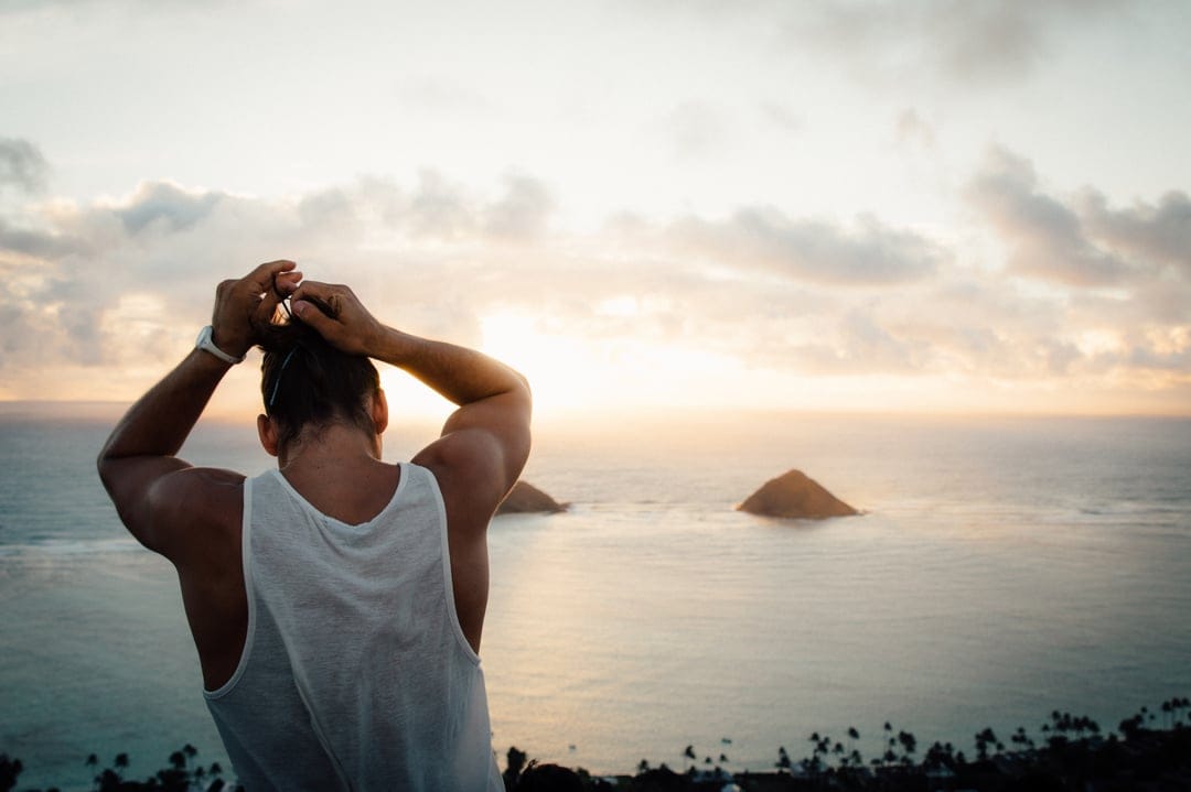 Man tying long hair from behind overlooks a sunset ocean with tropical islands and foreground palm leaves.