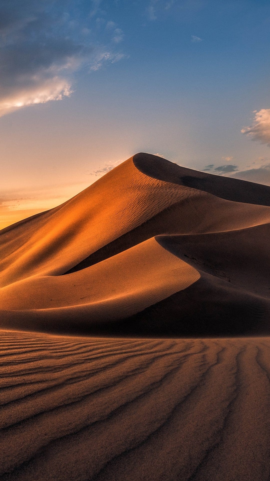 Rolling sand dunes with sharp ridges and deep shadows under a blue and orange sunset sky in a vast desert.