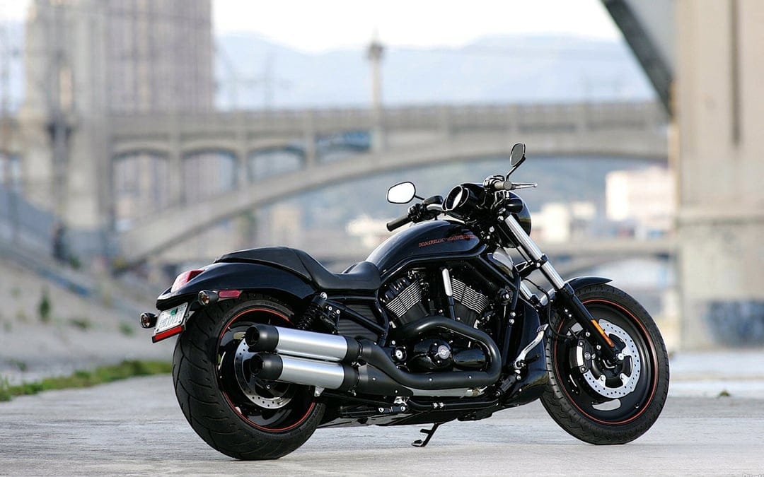 Black Harley-Davidson Night Rod motorcycle with silver exhaust on concrete against a blurred bridge.