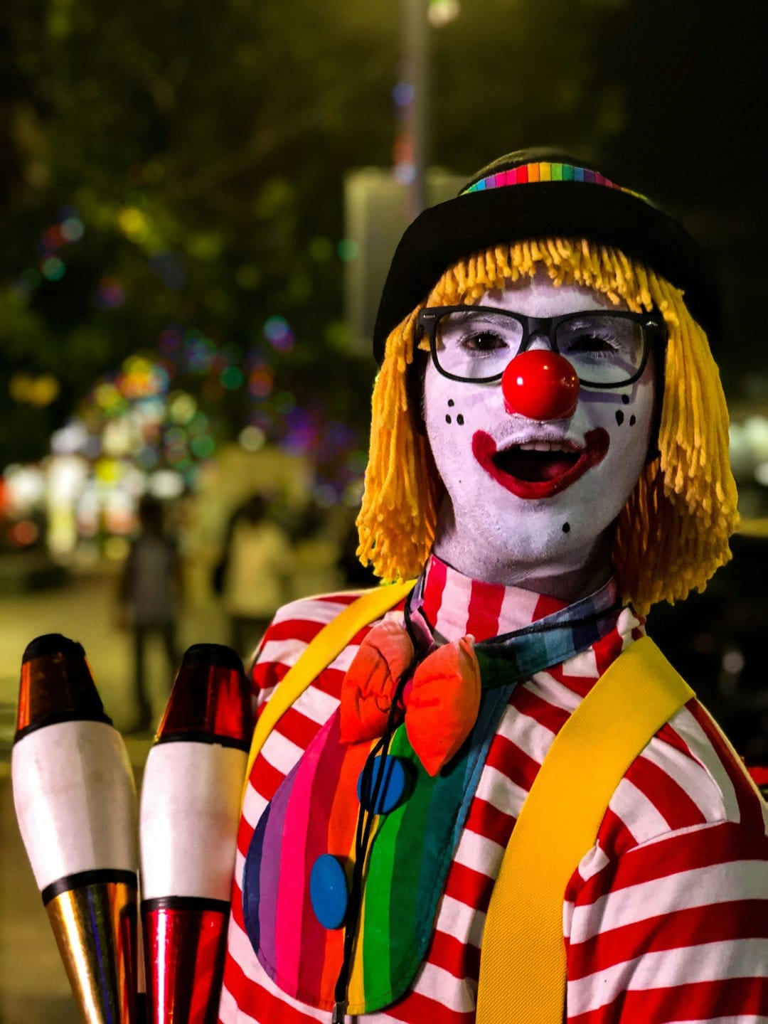Juggling clown with a yellow wig, red nose, and rainbow jabot holds pins against a dark carnival background.